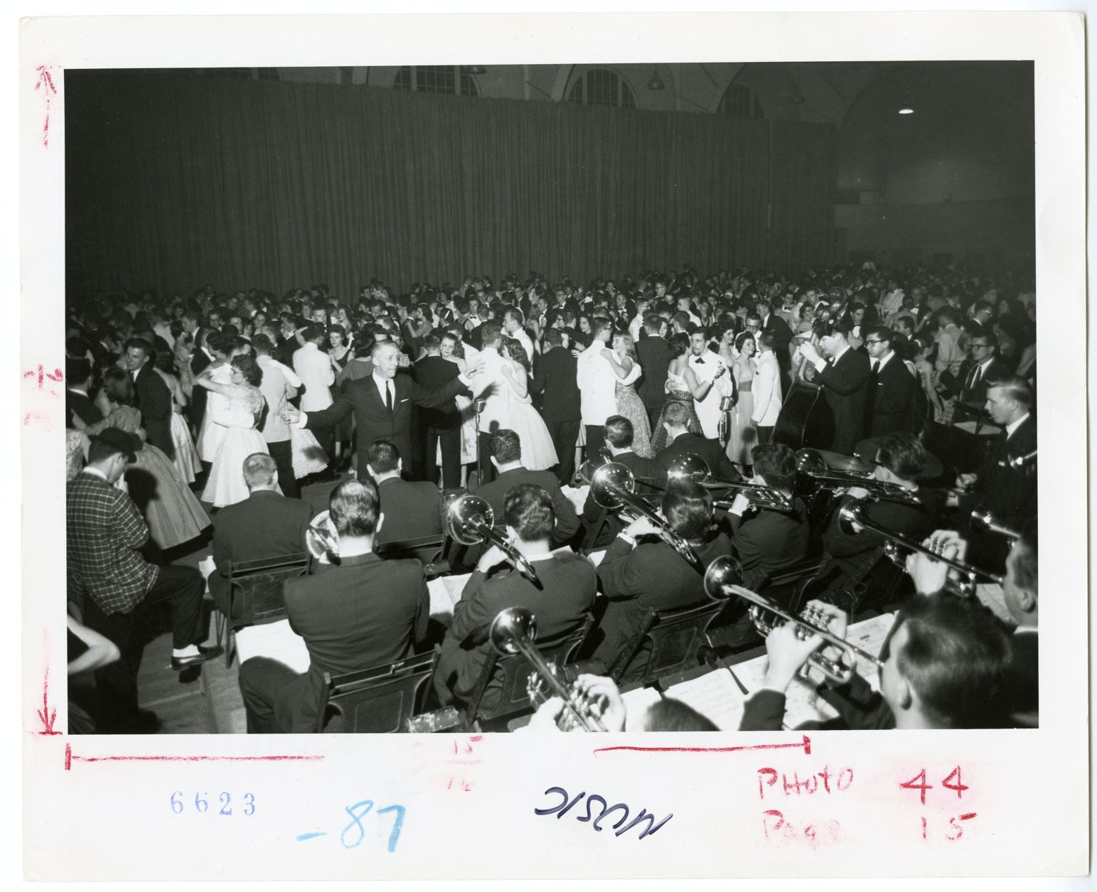 A view from behind the Stan Kenton Band as they perform in a gymnasium full of dancing college students, during Spring Formal. Written upside down on the front of the photograph: "Music." Written on the back of the photograph: "Spring Formal 1959, Stan Kenton Band," and typed on a sticker: "Music: instrumental."